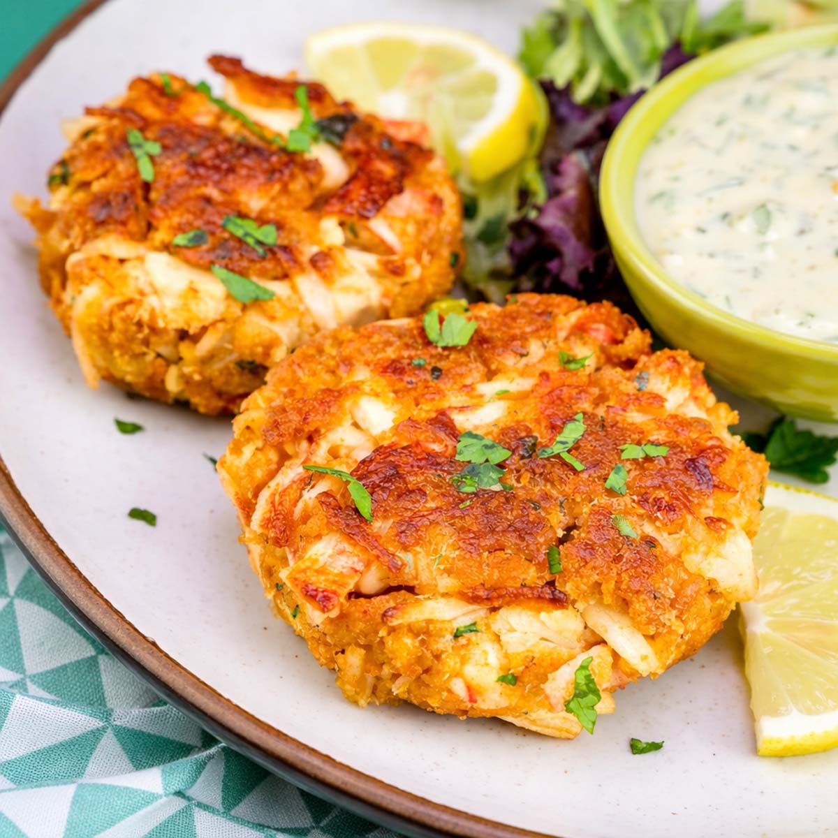 Two golden canned crab cakes on a plate with chopped parsley, lemon wedges, mixed greens, and a bowl of creamy dipping sauce. 