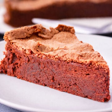 Close-up slice of flourless chocolate cake with a cracked, airy top and dense fudgy center on a white plate.