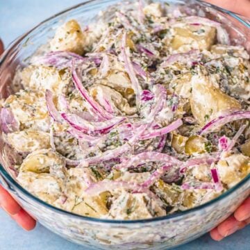 Creamy potato salad without eggs in a glass bowl with red onion slices, fresh dill, and tender potatoes coated in a rich herb dressing.