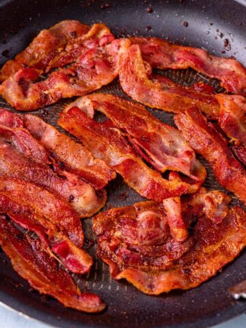 Crispy bacon cooking in a skillet on the stovetop, showing golden brown strips frying in their own rendered fat.