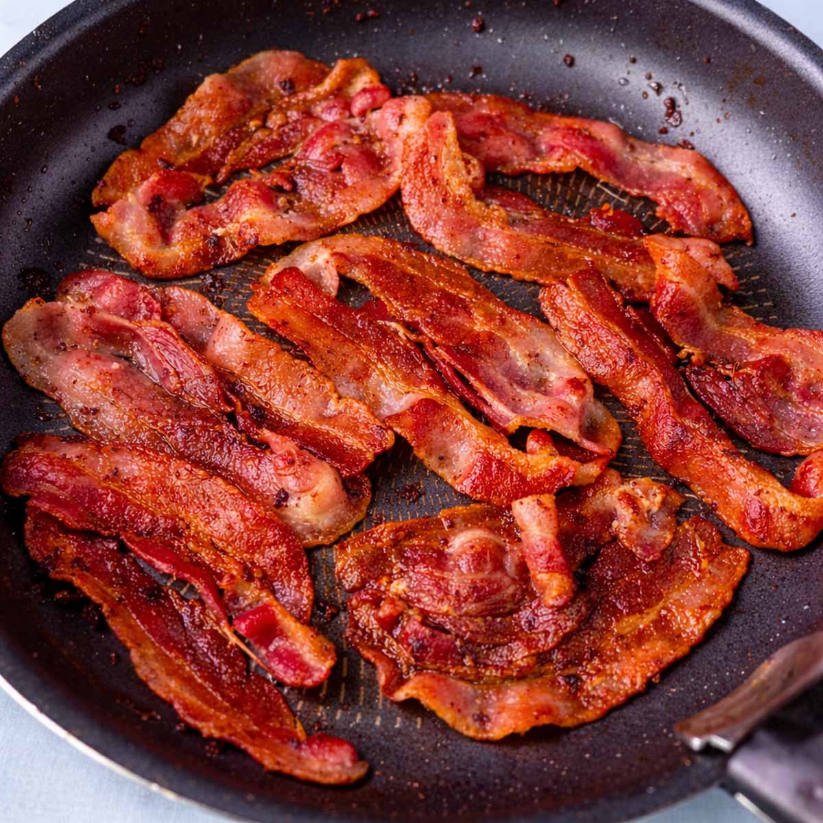 Crispy bacon cooking in a skillet on the stovetop, showing golden brown strips frying in their own rendered fat.