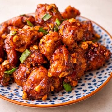 Korean fried chicken on a patterned plate, coated in a glossy spicy glaze and topped with chopped peanuts and green onions