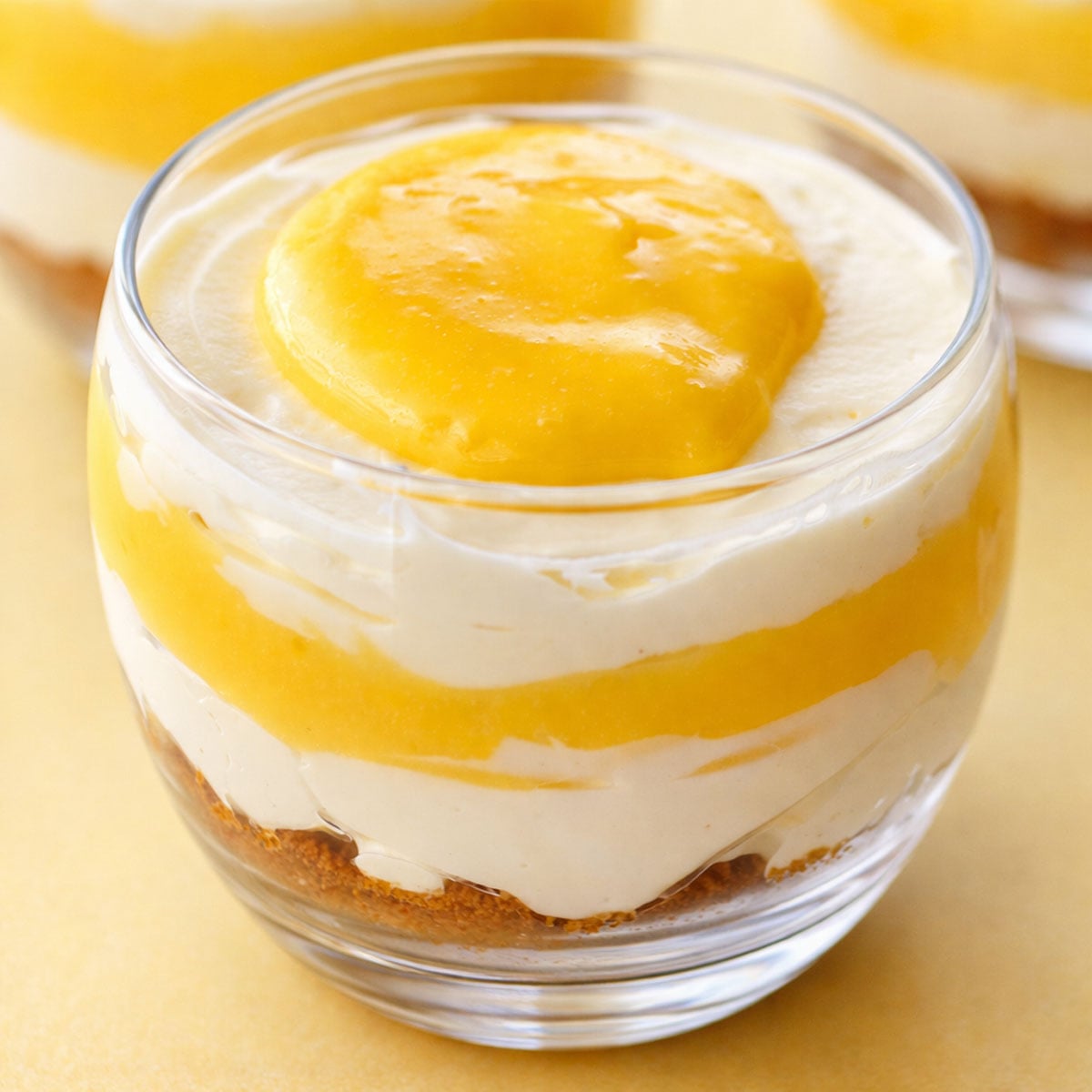 Lemon curd dessert cups with layered cheesecake filling and biscuit base in a glass, topped with glossy lemon curd