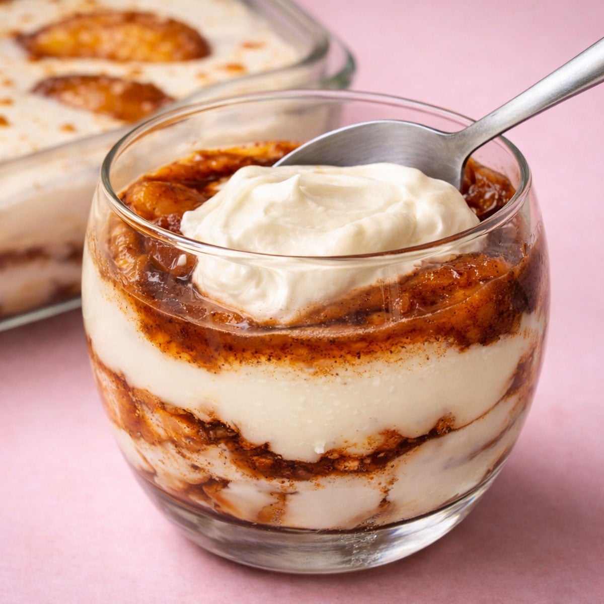 Peach cobbler pudding in a glass with creamy vanilla custard layers, cinnamon-spiced peaches, and a dollop of whipped cream with a spoon.