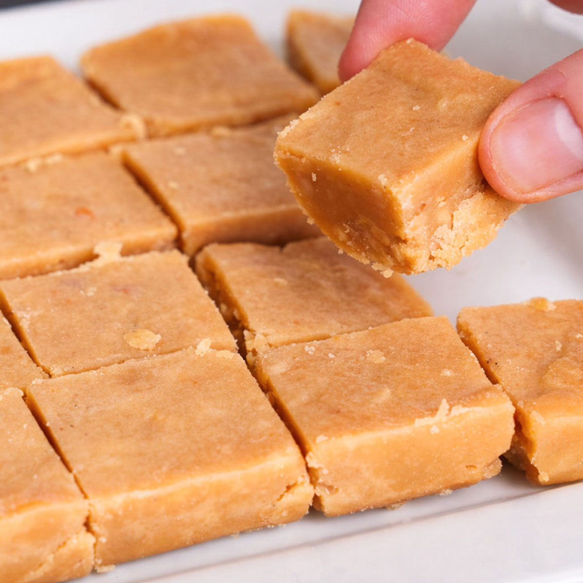 easy peanut butter fudge squares on a white plate with a hand lifting one piece, showing the smooth, creamy texture and soft, slightly crumbly center