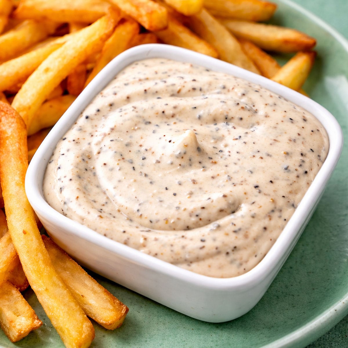 Close-up of creamy pepper mayonnaise in a white bowl served with crispy golden fries on a green plate