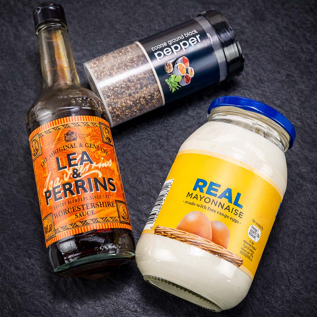 Ingredients for pepper mayonnaise: mayonnaise jar, ground black pepper grinder, and Worcestershire sauce bottle on a dark surface. 