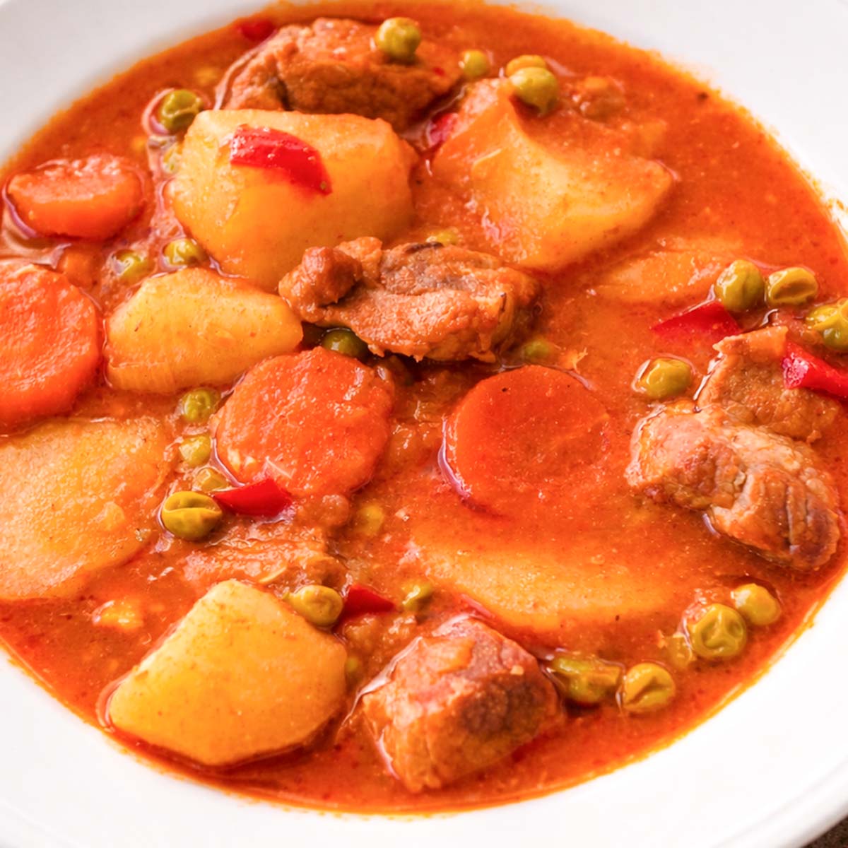 Spanish pork and potato stew with tender pork, carrots, peas, and red peppers in a rich tomato-based sauce served in a white bowl. 