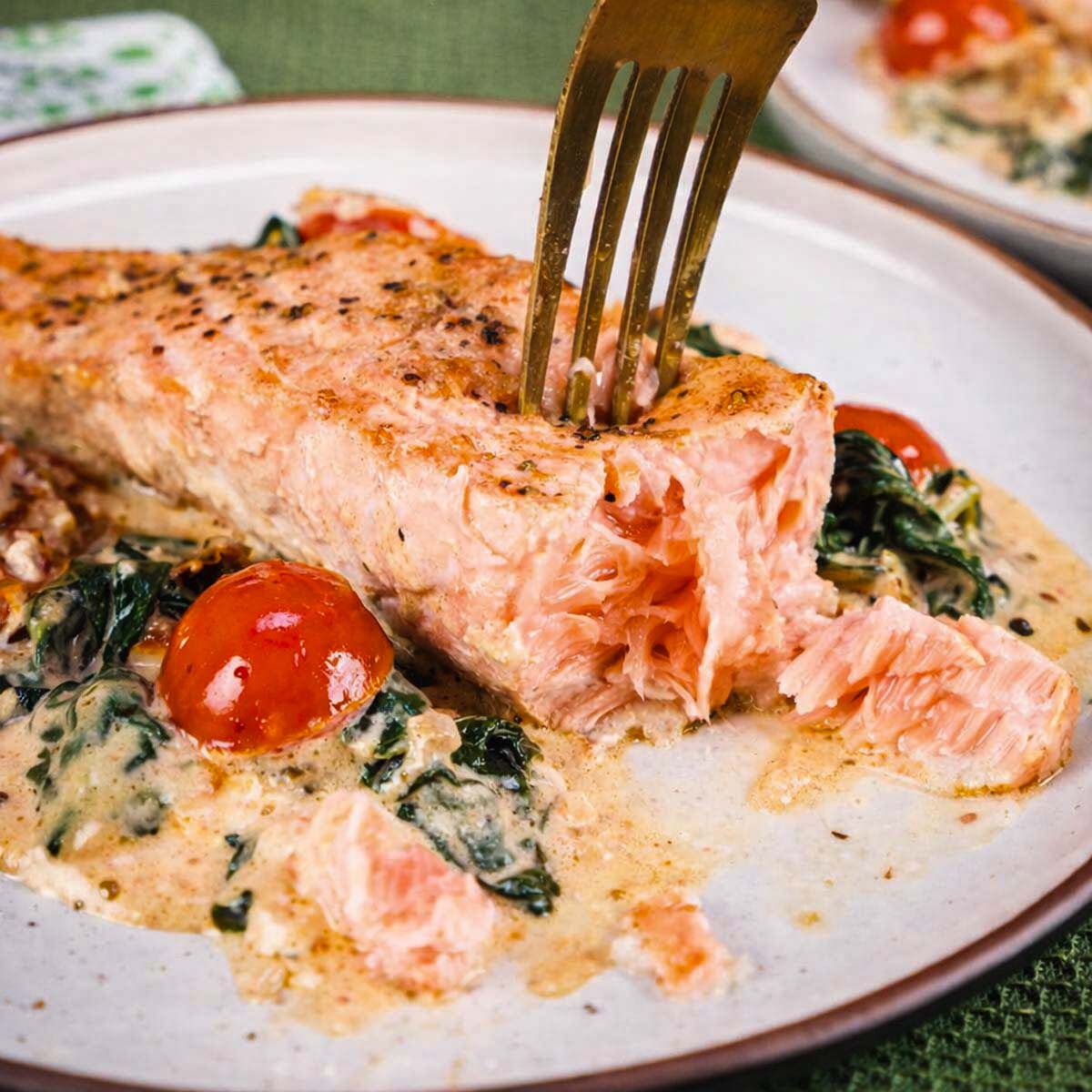 Fork flaking a salmon fillet served over creamy spinach sauce with cherry tomatoes on a white plate. 