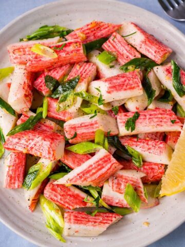 Sauteed imitation crab salad with green onions, parsley, and lemon wedge on a white plate, lightly seasoned and served fresh