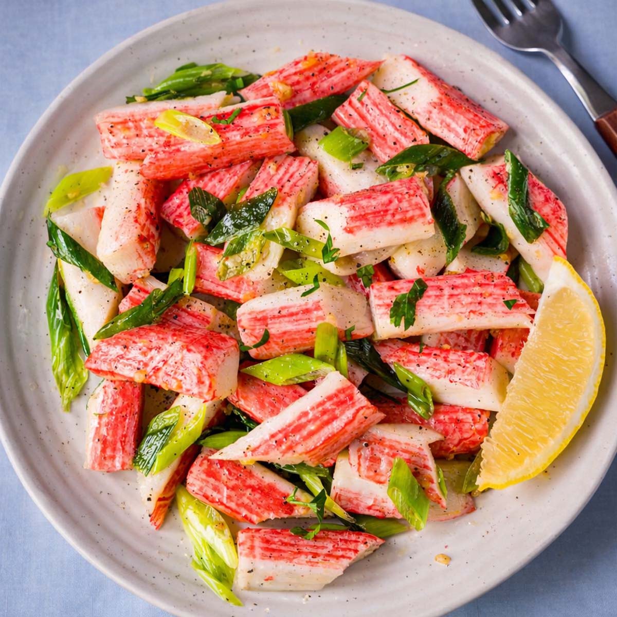 Sauteed imitation crab salad with green onions, parsley, and lemon wedge on a white plate, lightly seasoned and served fresh