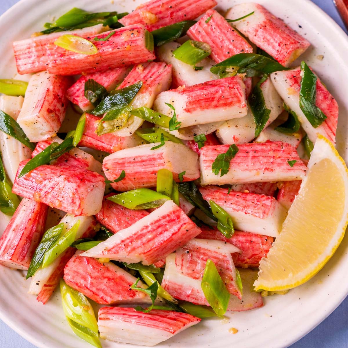 Sauteed imitation crab with garlic butter, green onions, and fresh herbs, served with a lemon wedge on a white plate