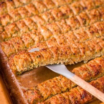 Sheet pan ground chicken kebabs sliced into strips on a baking tray with one piece lifted by a spatula, golden and slightly crisp on top.
