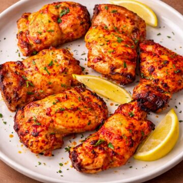 Tomato marinated chicken thighs baked until golden and slightly charred, served on a white plate with lemon wedges and fresh herbs