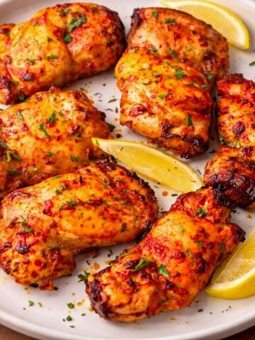 Tomato marinated chicken thighs baked until golden and slightly charred, served on a white plate with lemon wedges and fresh herbs