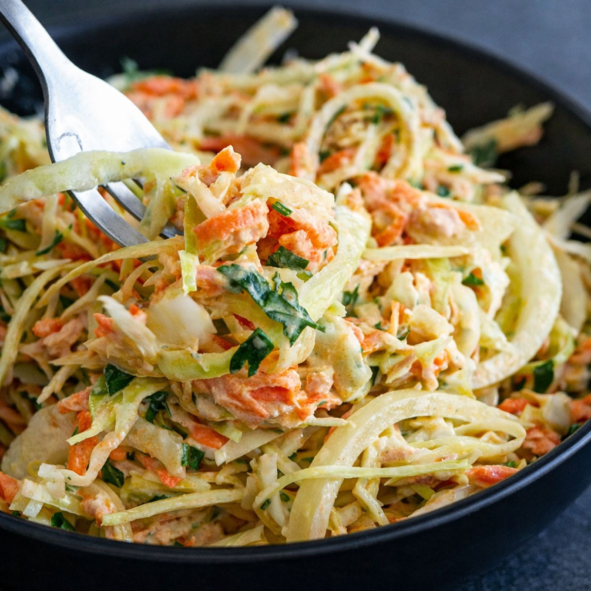 Fork lifting a bite of creamy tuna cabbage salad with shredded cabbage, carrots, onion, and herbs in a black bowl. 