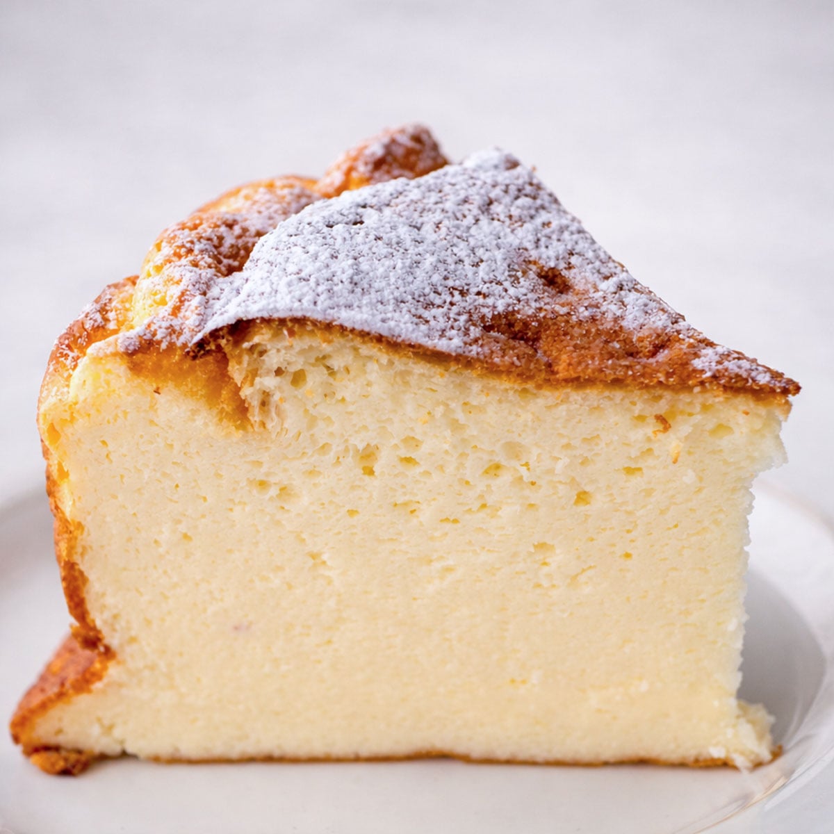 Close-up of a thick slice of easy yogurt cake with a golden top dusted in powdered sugar, showing a light, airy crumb on a white plate. 