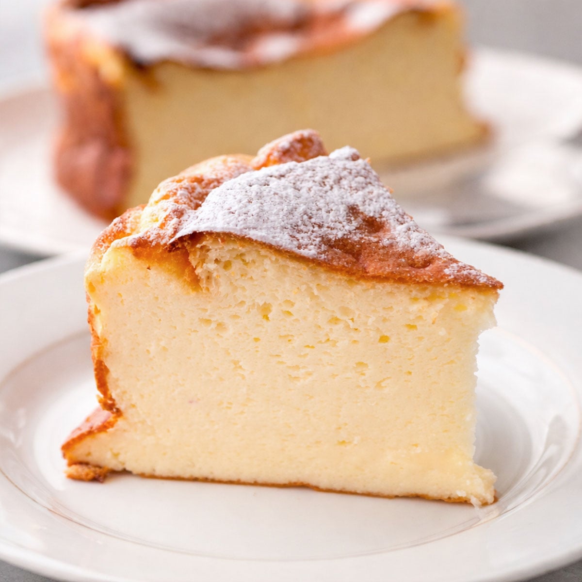 Slice of easy yogurt cake on a white plate, topped with powdered sugar and a golden-brown crust, with the rest of the cake softly blurred in the background. 