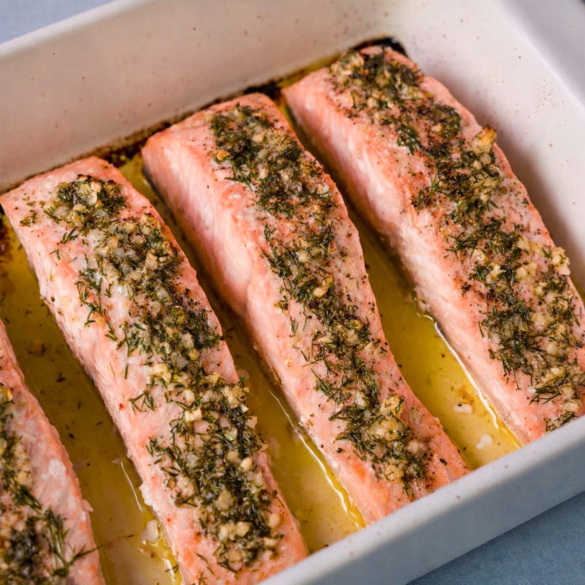 close up baked lemon dill salmon fillets in a white baking dish with garlic, fresh dill, and olive oil juices