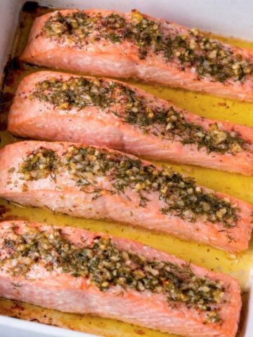 baked lemon dill salmon fillets in a white baking dish with garlic, fresh dill, and olive oil juices