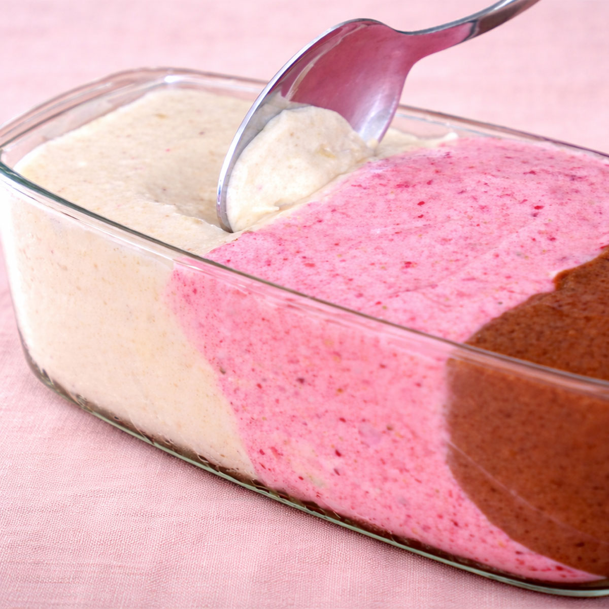 Close up banana nice cream in a glass loaf pan with vanilla, strawberry, and chocolate layers on soft pink linen background