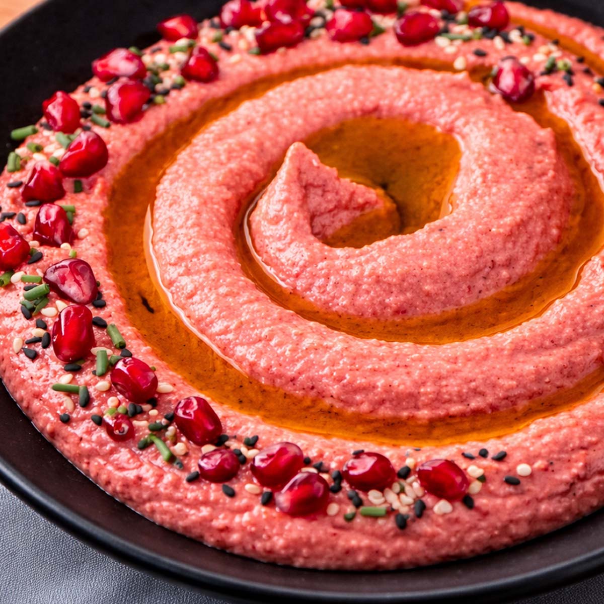 Close-up of creamy beet hummus swirled with olive oil and topped with pomegranate seeds and sesame in a black bowl