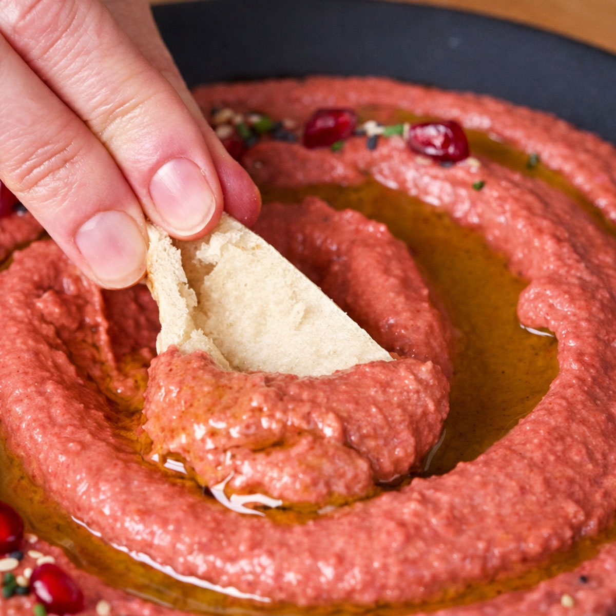 Hand dipping pita bread into smooth easy beet hummus dip with olive oil and pomegranate seeds