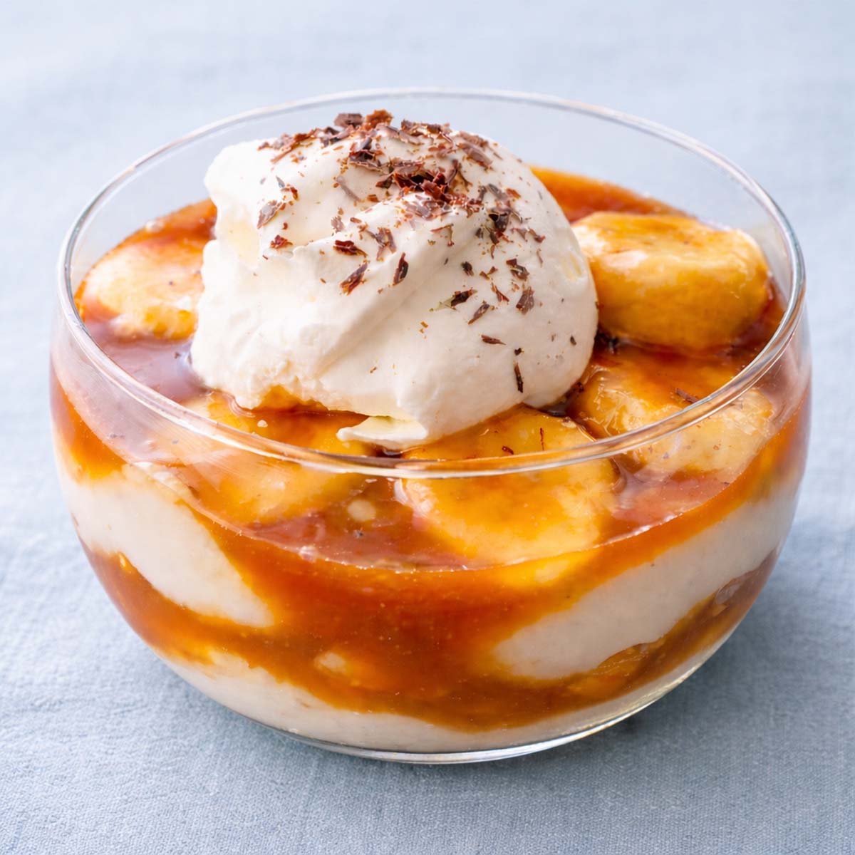 banana caramel pudding layered with vanilla custard and sliced bananas topped with whipped cream and chocolate shavings in a glass dessert cup