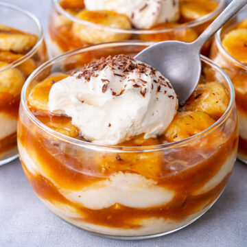 banana caramel pudding with vanilla custard and sliced bananas topped with whipped cream and chocolate shavings in a glass dessert bowl