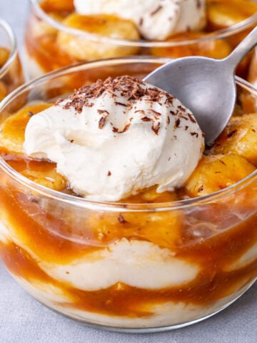 banana caramel pudding with vanilla custard and sliced bananas topped with whipped cream and chocolate shavings in a glass dessert bowl