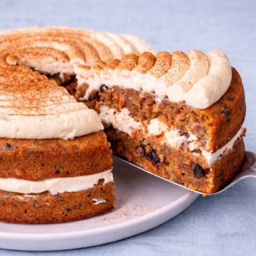 Carrot cake layered with cream cheese frosting and cinnamon on top, with a slice cut out showing the moist carrot and raisin filling.