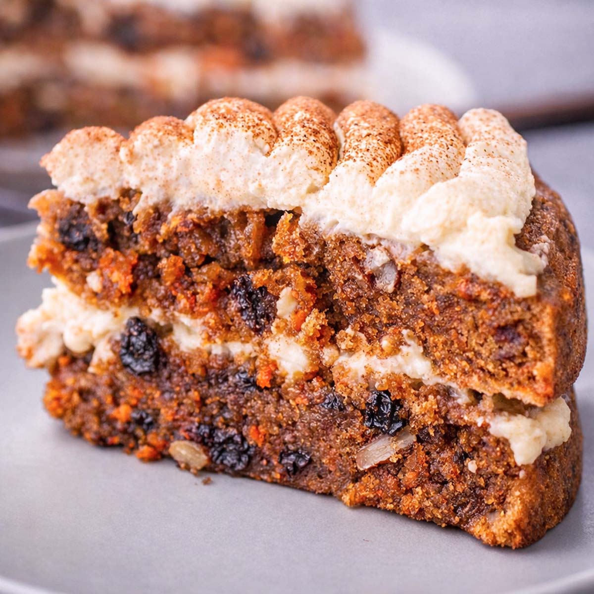 Slice of moist carrot cake with cream cheese frosting, raisins, walnuts, and grated carrots, topped with piped frosting and cinnamon on a gray plate.