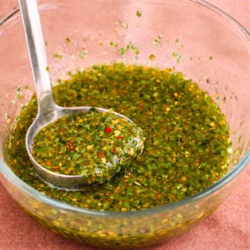 Homemade chimichurri sauce in a glass bowl with a ladle, showing fresh parsley, garlic, olive oil, and red pepper flakes on a terracotta linen background.