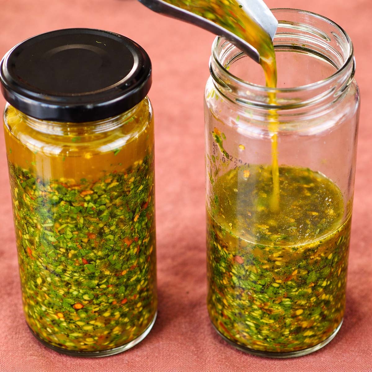 Pouring homemade lemon chimichurri sauce into a glass jar beside another jar filled with fresh chimichurri sauce on a terracotta linen background.