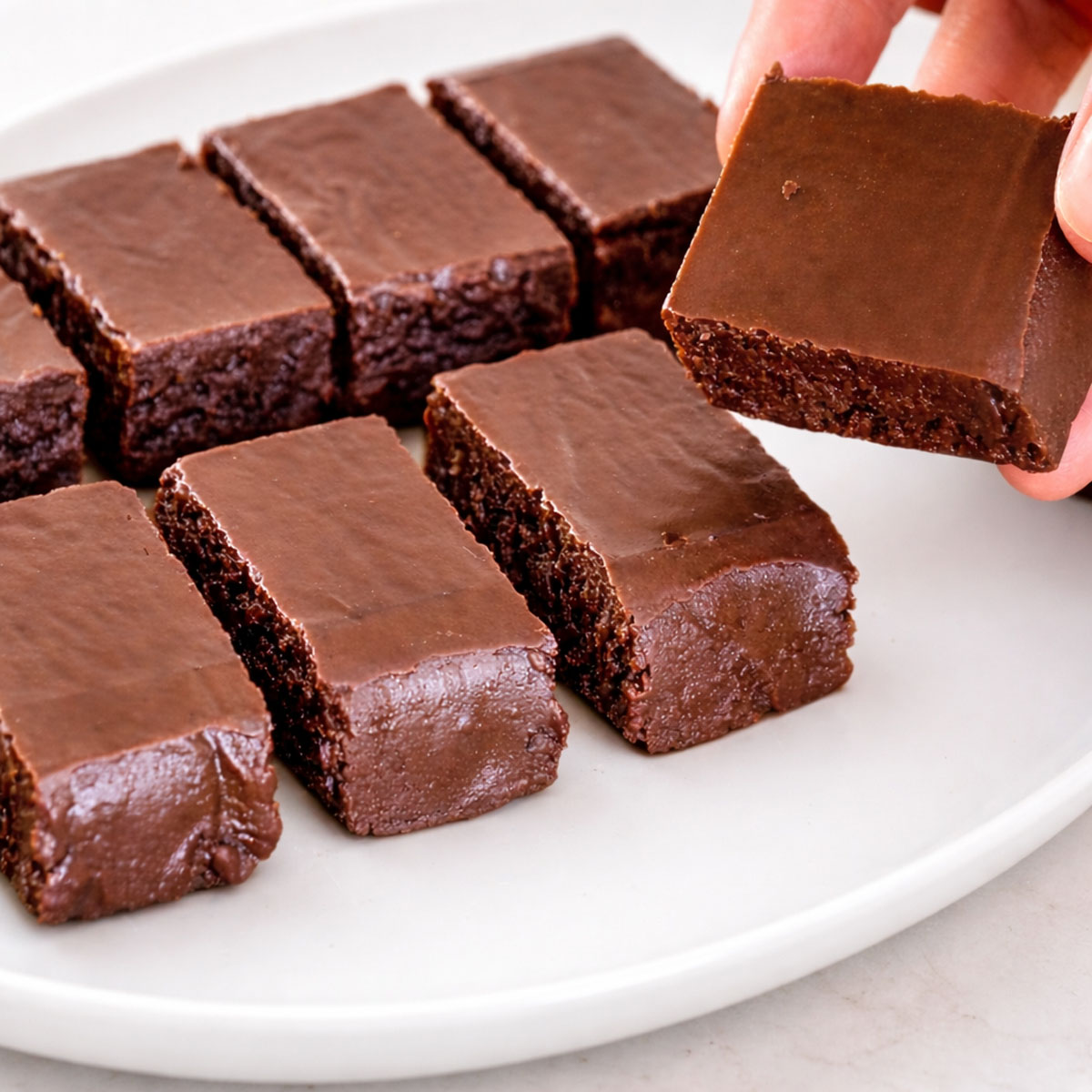 Hand lifting a piece of chocolate caramels with a glossy chocolate top and soft caramel center from a white plate.