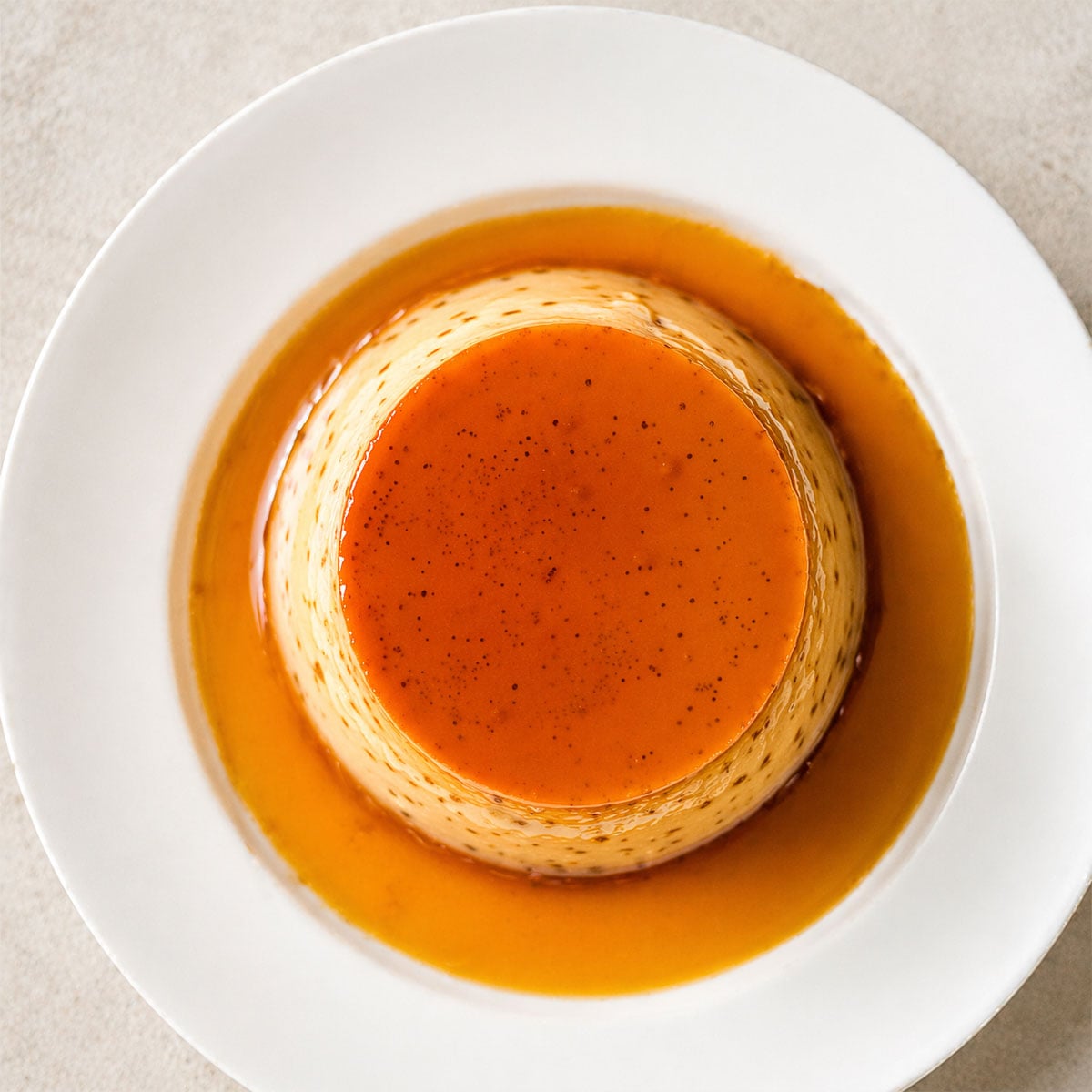 Top-down view of coffee flan with glossy caramel sauce on a white plate over warm linen background