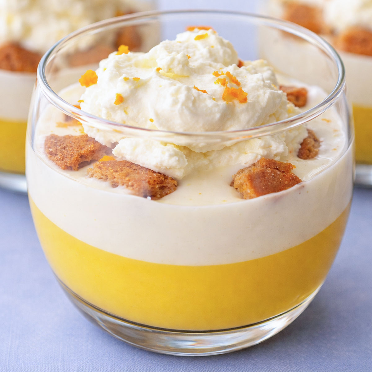 Creamy orange pudding in a glass dessert cup layered with smooth citrus pudding, vanilla cream, biscuit pieces, and whipped cream topped with fresh orange zest.