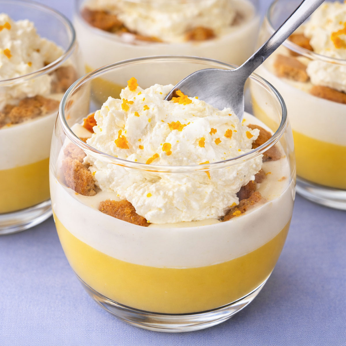 Creamy orange pudding dessert in a glass cup with layers of citrus pudding, vanilla cream, biscuit pieces, and whipped cream topped with orange zest and a spoon.
