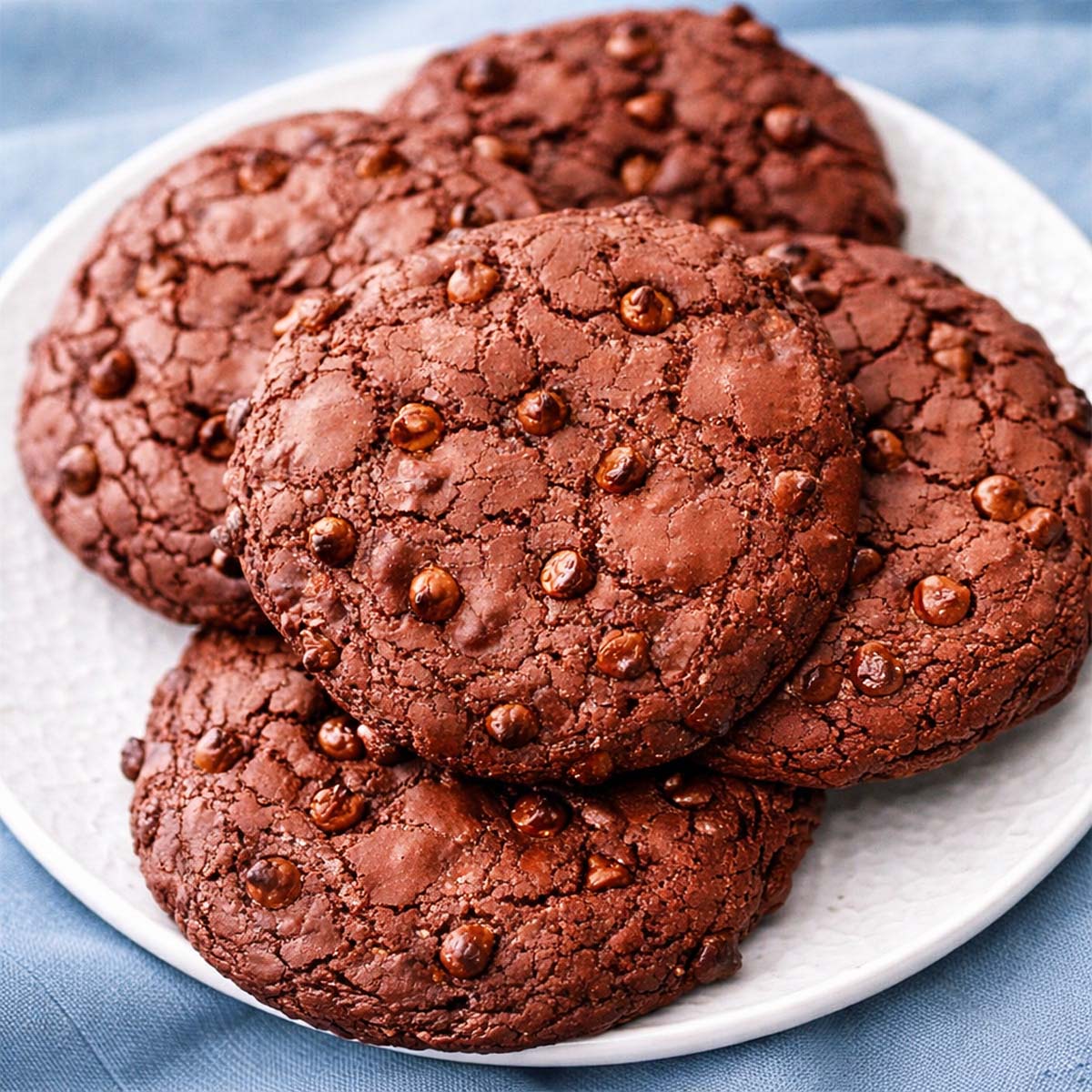Double chocolate chip cookies stacked on a white plate with a soft blue linen background, showing rich chocolate texture and chocolate chips on top.