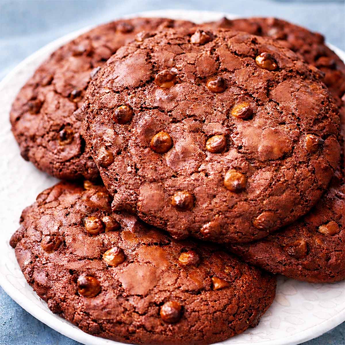 Double chocolate chip cookies on a white plate with a soft blue linen background, showing thick cookies with cracked chocolate tops and chocolate chips throughout.