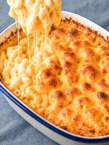 easy baked mac and cheese in a white casserole dish with golden bubbly cheese crust and creamy macaroni being lifted with melted cheese strings