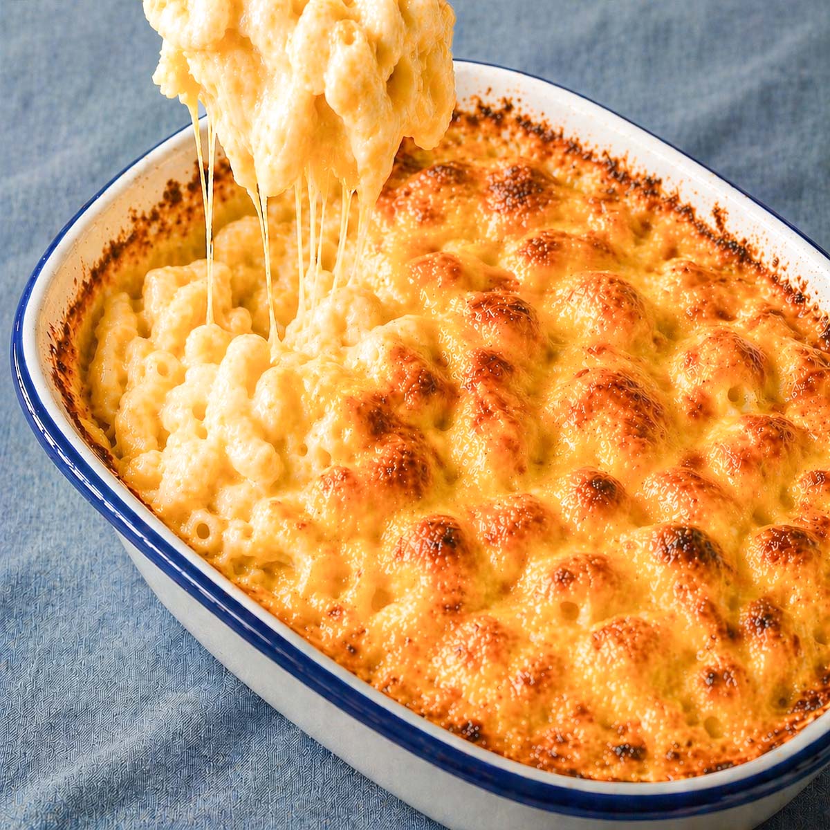 easy baked mac and cheese in a white casserole dish with golden bubbly cheese crust and creamy macaroni being lifted with melted cheese strings