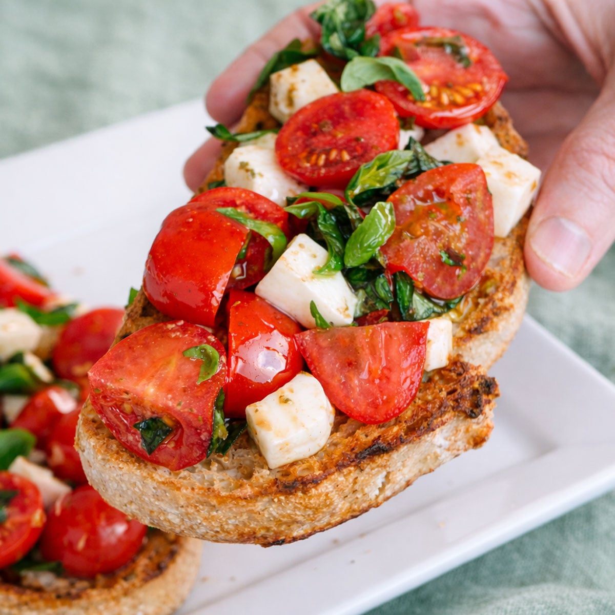 Hand holding a slice of cherry tomato bruschetta topped with mozzarella, basil, and olive oil on toasted bread.