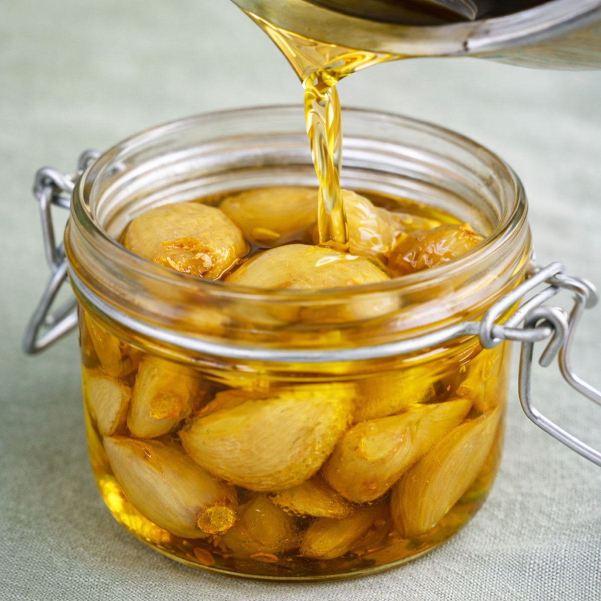 Garlic confit in a glass jar with golden olive oil being poured over soft cooked garlic cloves on a soft sage linen background
