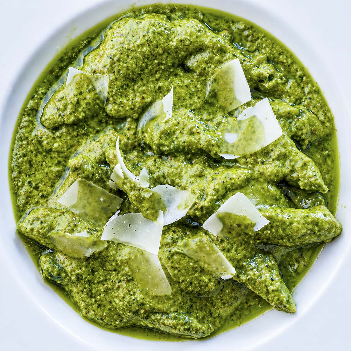 Fresh basil pesto pasta topped with shaved Parmesan cheese in a white bowl, close-up overhead view