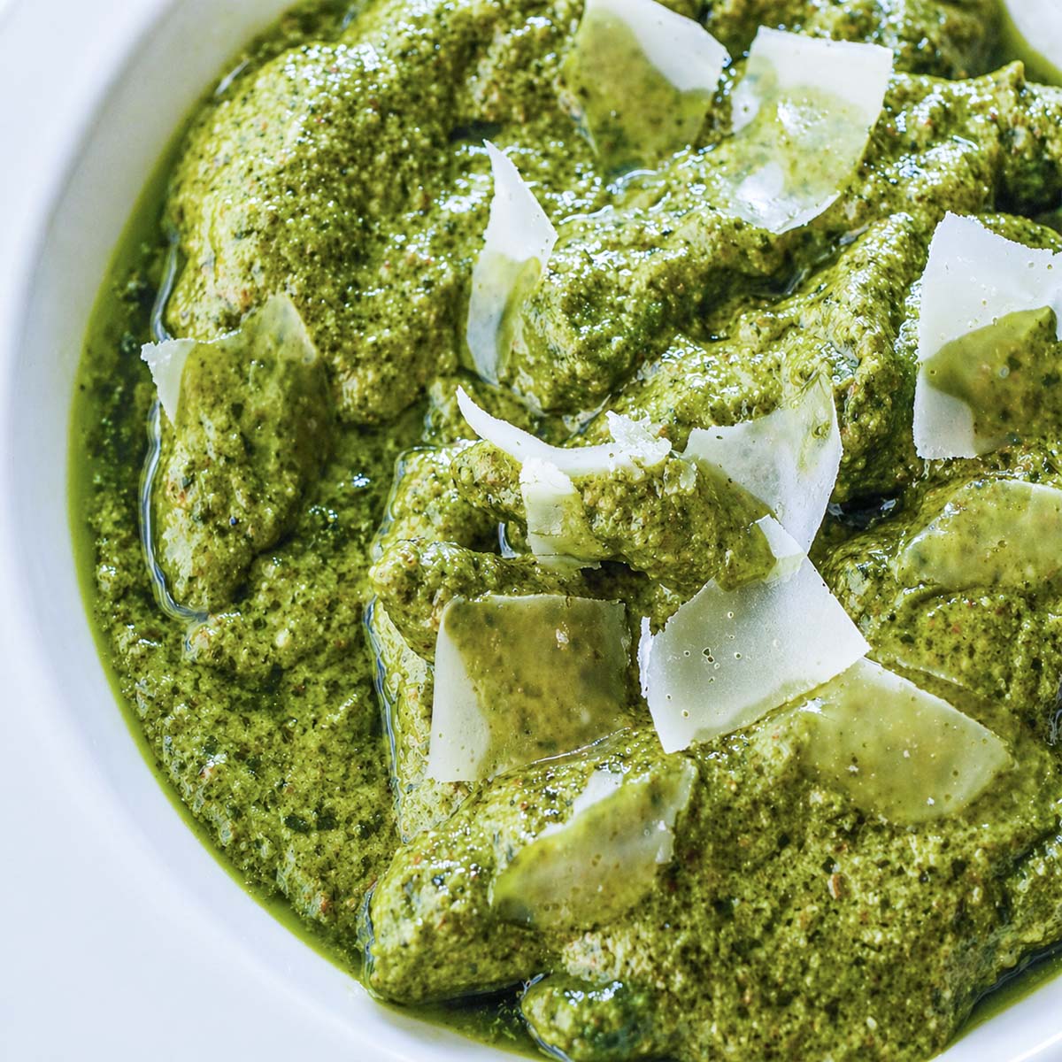 Close-up of fresh basil pesto pasta with shaved Parmesan cheese in a white bowl, coated in rich green basil pesto sauce