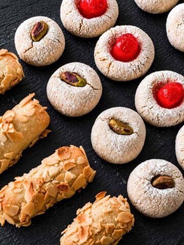 Italian almond cookies made with almond flour on a black slate board, including amaretti-style cookies with cherries, pistachios, and almonds.
