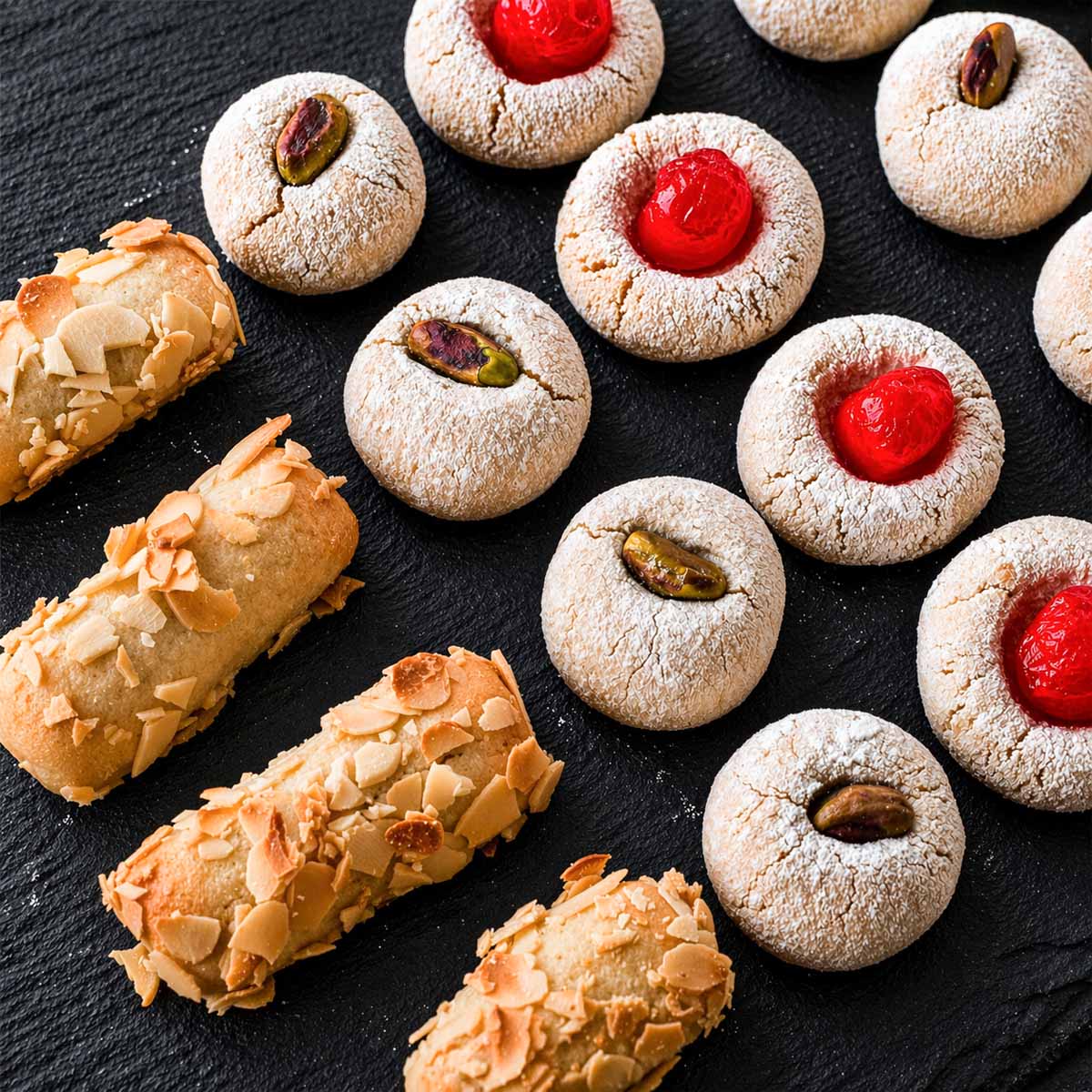 Italian almond cookies made with almond flour on a black slate board, including amaretti-style cookies with cherries, pistachios, and almonds.