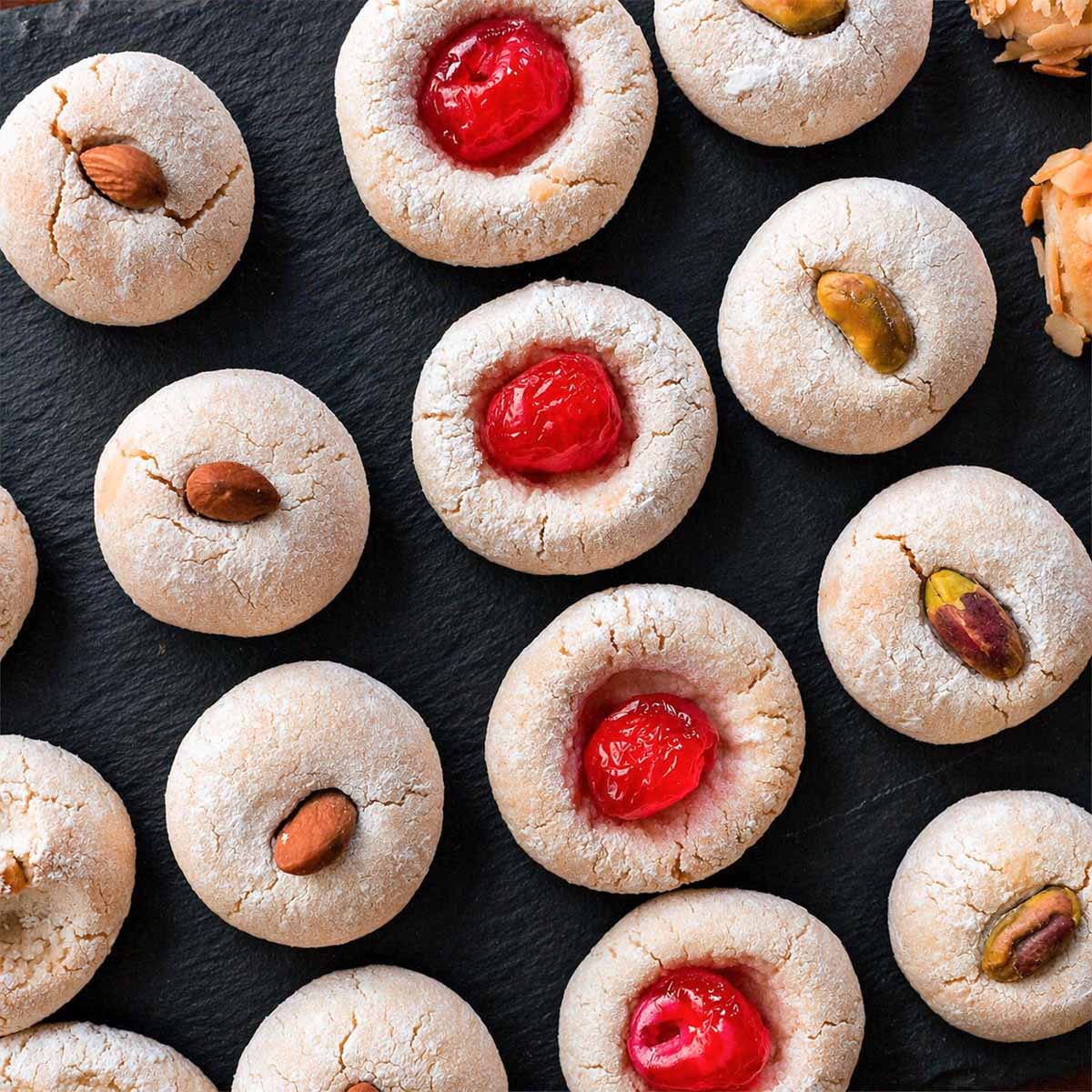 Italian almond cookies made with almond flour arranged on a black slate board, topped with cherries, almonds, and pistachios.