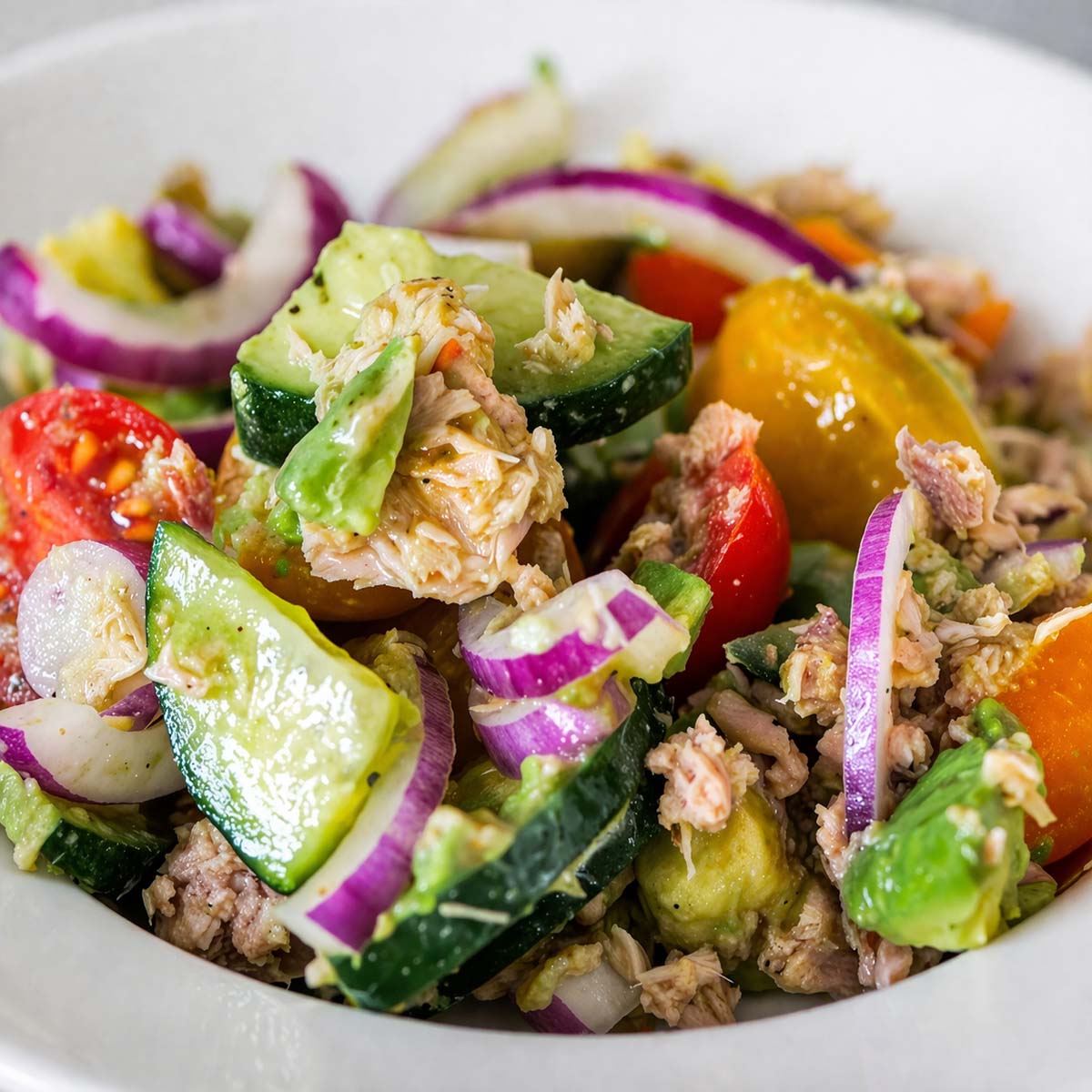 Mediterranean tuna salad with avocado, cucumber, cherry tomatoes, and red onion in a white bowl, close-up view with light dressing